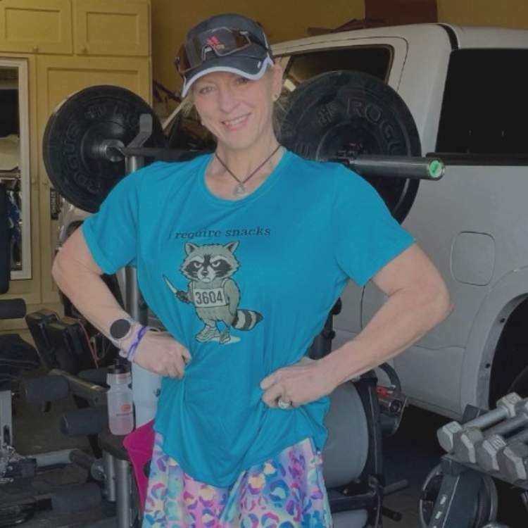 A woman wearing a blue t-shirt featuring a cartoon grey raccoon wearing a "3604" bib, holding a small knife, and the text "i require snacks", 