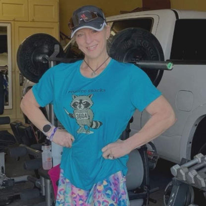 A woman wearing a blue t-shirt featuring a cartoon grey raccoon wearing a "3604" bib, holding a small knife, and the text "i require snacks", 