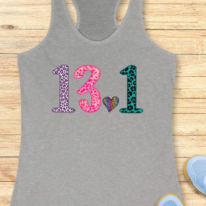 A heather gray racerback tank top laid flat on a light wood plank background, featuring the number '13.1' in leopard-print style with a small multicolor heart in the center.