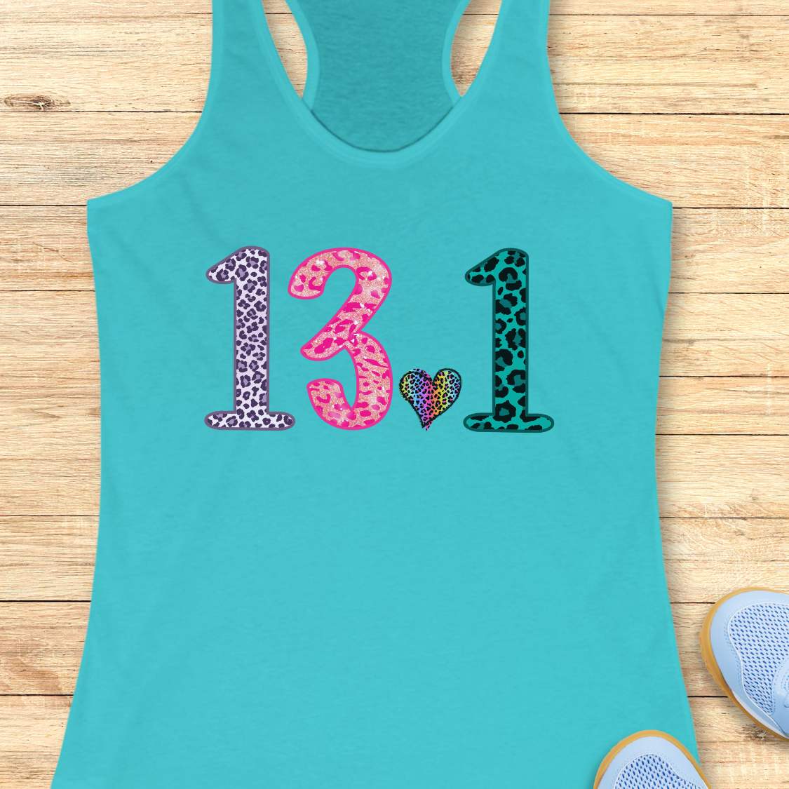 A turquoise racerback tank top laid flat on a light wood background, featuring the number '13.1' and a small heart in colorful leopard-print patterns across the chest.