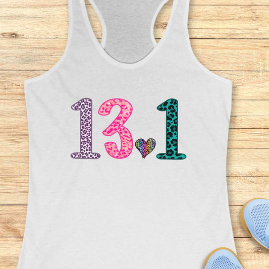 A white racerback tank top laid flat on a light wood background, featuring the numbers '13.1' in leopard print patterns (purple, pink, and teal) with a small multicolor heart between them.