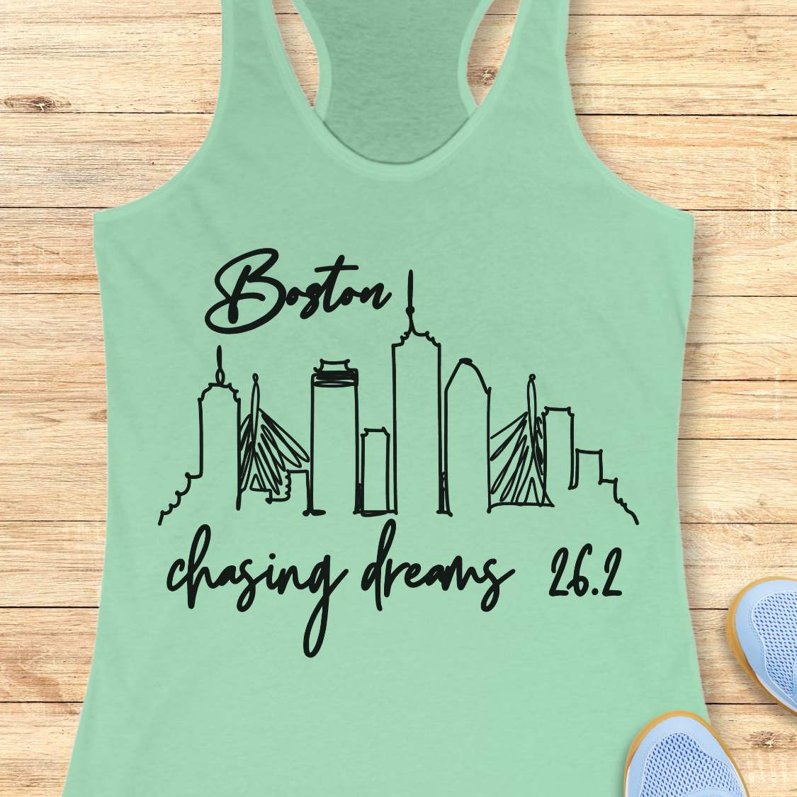 Light green tank top with black graphic design of Boston skyline and text Boston chasing dreams 26.2, on a wooden surface