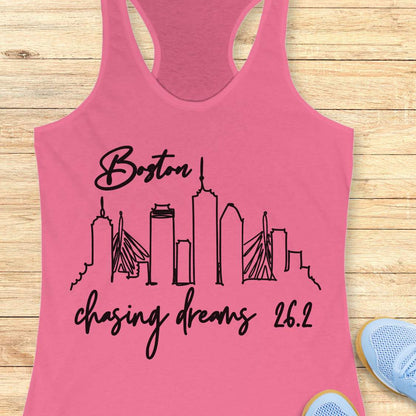 Pink tank top with black graphic design of Boston skyline and text Boston chasing dreams 26.2, on a wooden surface