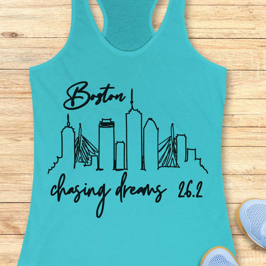 Turquoise tank top with black skyline design and text Boston chasing dreams 26.2, on a wooden surface