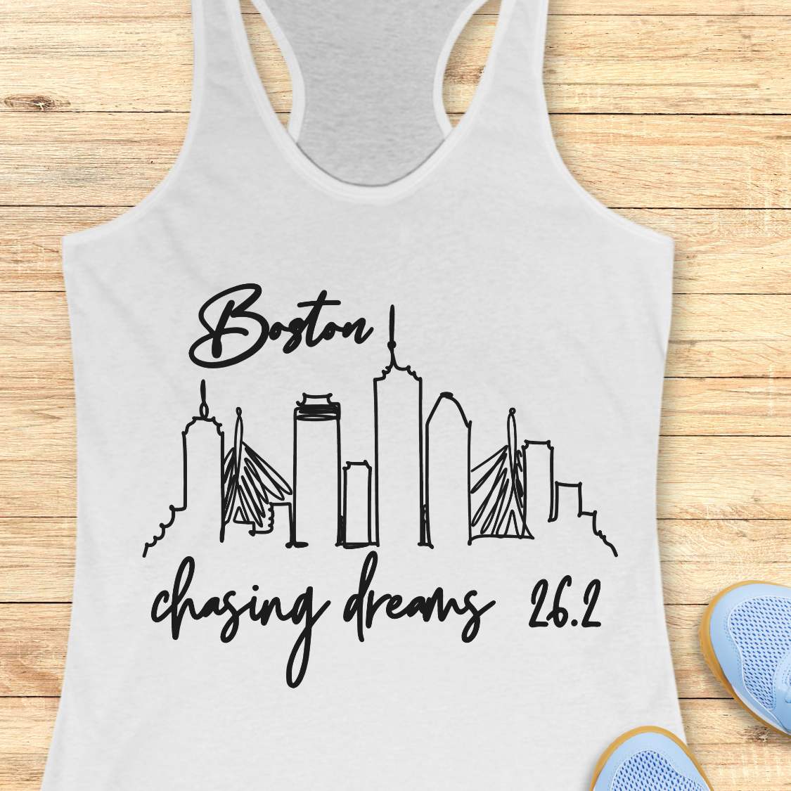 white tank top with Boston skyline and marathon text Boston chasing dreams 26.2, on a wooden surface