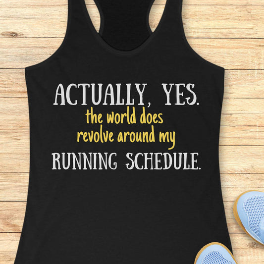 Black tank top with humorous text 'Actually, yes. the world does revolve around my running schedule.' on a wooden surface