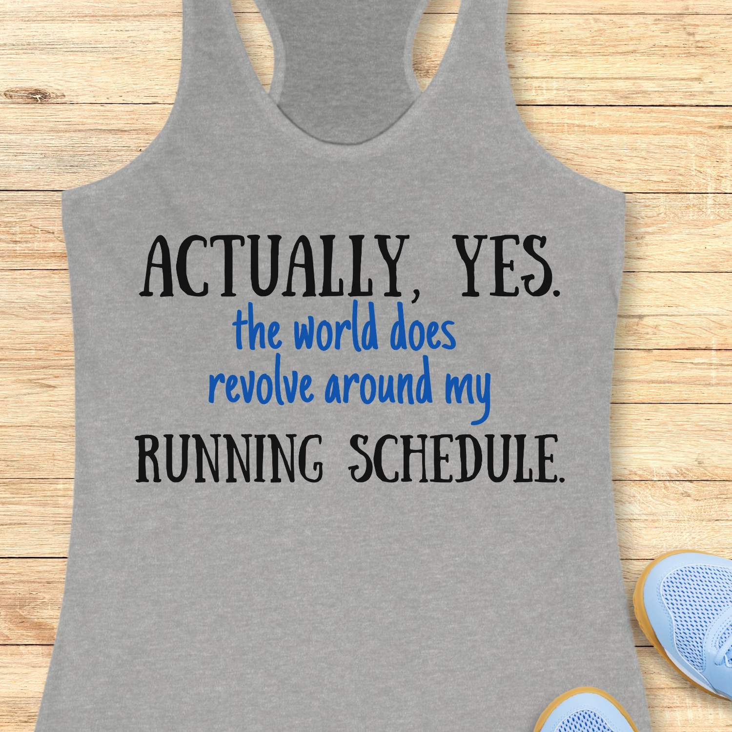 Gray tank top with humorous text 'Actually, yes. the world does revolve around my running schedule.' on a wooden surface