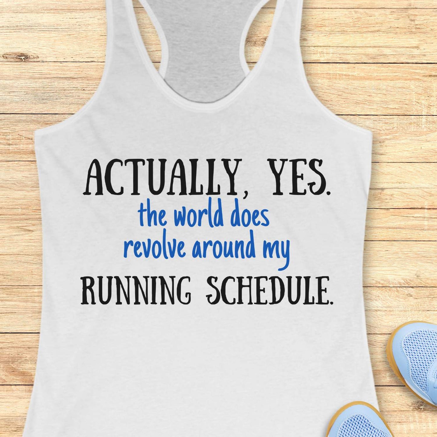white tank top with humorous text 'Actually, yes. the world does revolve around my running schedule.' on a wooden surface