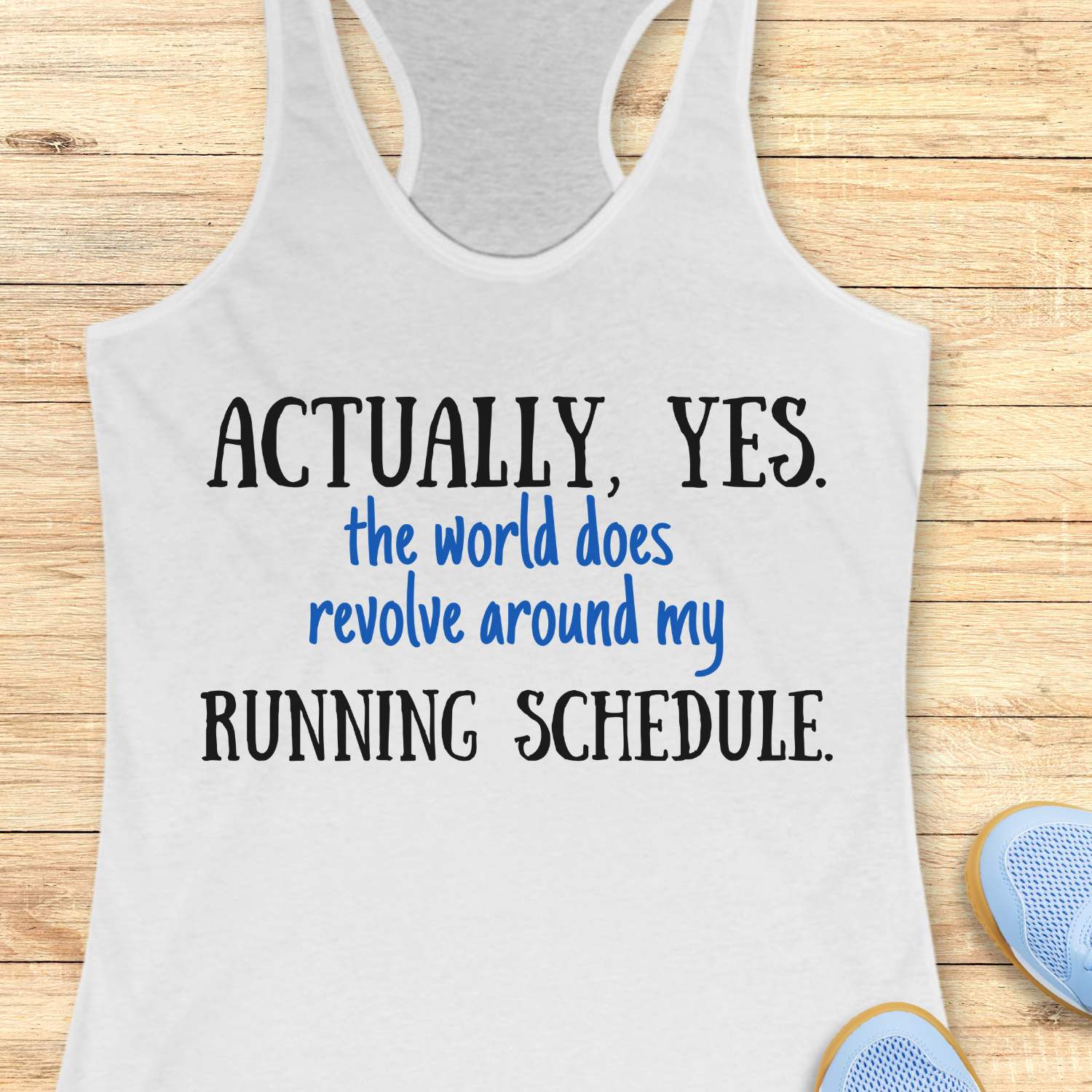 white tank top with humorous text 'Actually, yes. the world does revolve around my running schedule.' on a wooden surface