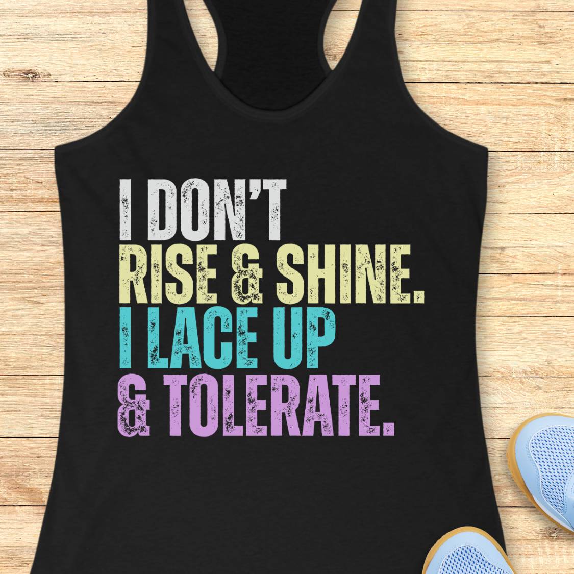 A black racerback tank top laid flat on a light wood surface, featuring the motivational text 'I DON'T RISE & SHINE. I LACE UP & TOLERATE.