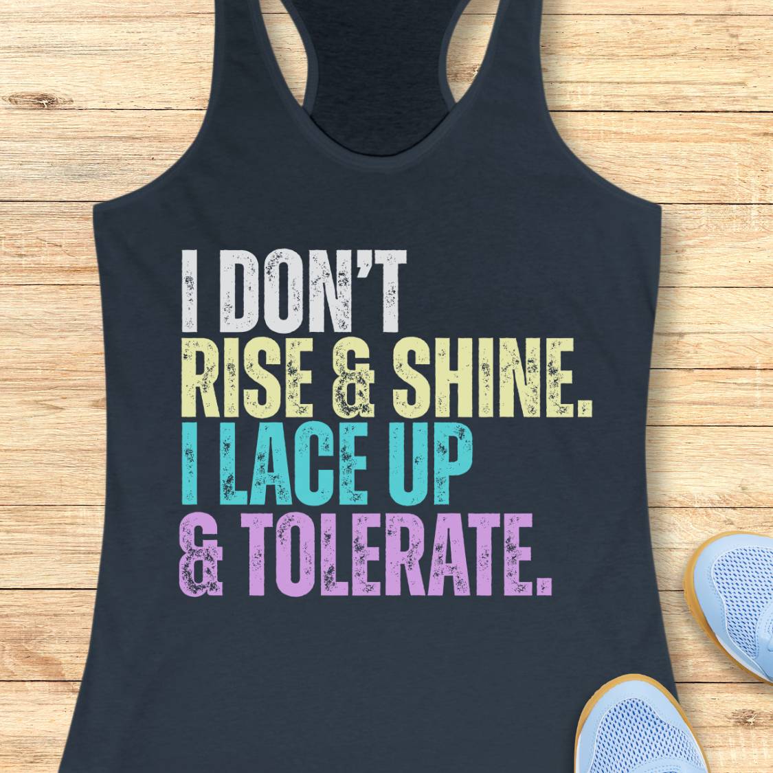 An indigo racerback tank top laid flat on a light wood surface, featuring the motivational text 'I DON'T RISE & SHINE. I LACE UP & TOLERATE.