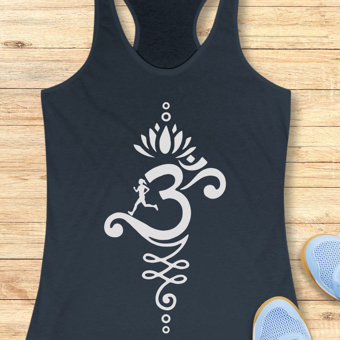 An indigo racerback tank top with a large white Om symbol graphic featuring a lotus flower and a small running figure, laid flat on a light wood background with blue sneakers partially visible at the bottom right.
