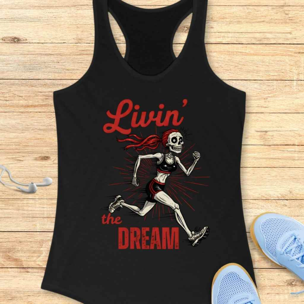 Graphic design on a black racerback tank top depicting a lively female skeleton runner with red flowing hair, wearing a black sports bra and shorts with red accents, surrounded by red sunburst lines and the red text Livin' the DREAM, displayed on a wooden surface with athletic accessories