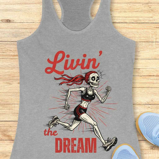 Graphic design on a grey tank top featuring a running female skeleton with a red ponytail, black sports bra and shorts, and red script text "Livin' the Dream" on a wooden background next to light blue athletic shoes