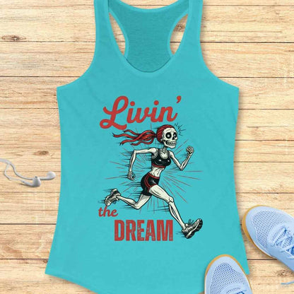 Turquoise racerback tank top displaying a cartoon running skeleton with a red ponytail, black sports bra and shorts, and the red text "Livin' the DREAM" with sunburst lines, laid on a light wooden surface with white earbuds and light blue running shoes nearby