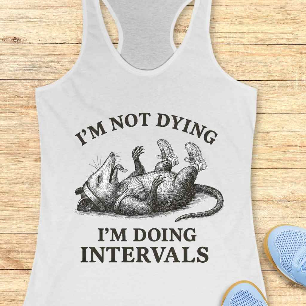 Graphic print on a white tank top features an opossum character lying on its back, appearing exhausted with its tongue out, wearing a small tank top, headband, and sneakers, above the motivational and humorous text I'M NOT DYING I'M DOING INTERVALS, presented on a light wood grain surface with blue running shoes visible at the bottom right