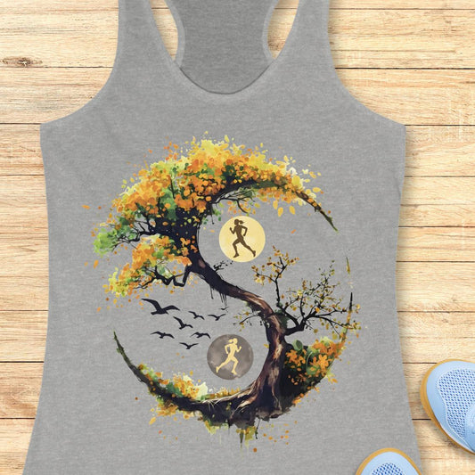 A grey racerback tank top laid flat on a light wood surface, featuring a large graphic of a stylized tree with autumn leaves in yellow, orange, and brown, integrated with a running yin-yang symbol showing two running figures and birds.