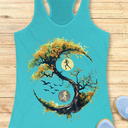 A turquoise racerback tank top laid flat on a light wood surface, featuring a large graphic of a stylized tree with autumn leaves in yellow, orange, and brown, integrated with a running yin-yang symbol showing two running figures and birds.