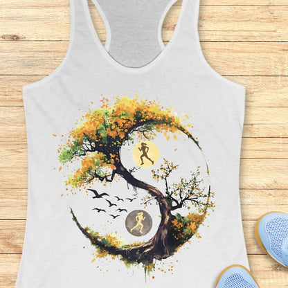 A white racerback tank top laid flat on a light wood surface, featuring a large graphic of a stylized tree with autumn leaves in yellow, orange, and brown, integrated with a running yin-yang symbol showing two running figures and birds.