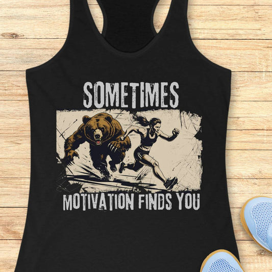 A black racerback tank top laid flat on a light wood surface, featuring a distressed graphic of a bear chasing a woman with the text 'SOMETIMES MOTIVATION FINDS YOU' in white. Part of a blue shoe is visible at the bottom right as a styling prop.
