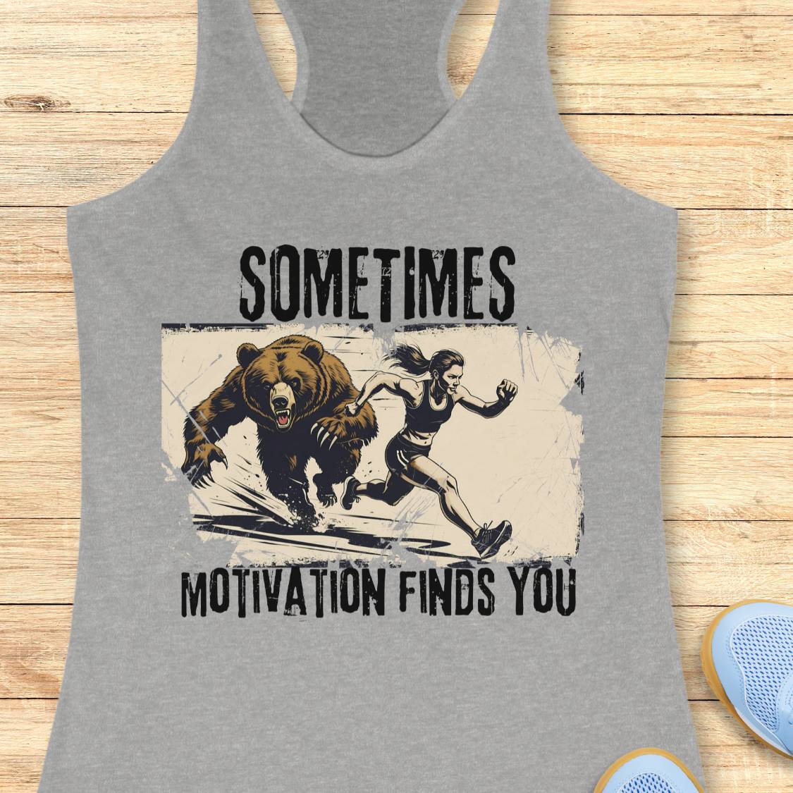 A heather gray racerback tank top laid flat on a light wood background, featuring a distressed graphic of a woman running from a bear with the text 'SOMETIMES MOTIVATION FINDS YOU'.