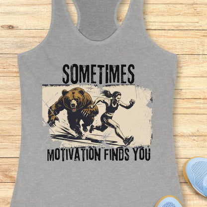 A heather gray racerback tank top laid flat on a light wood background, featuring a distressed graphic of a woman running from a bear with the text 'SOMETIMES MOTIVATION FINDS YOU'.
