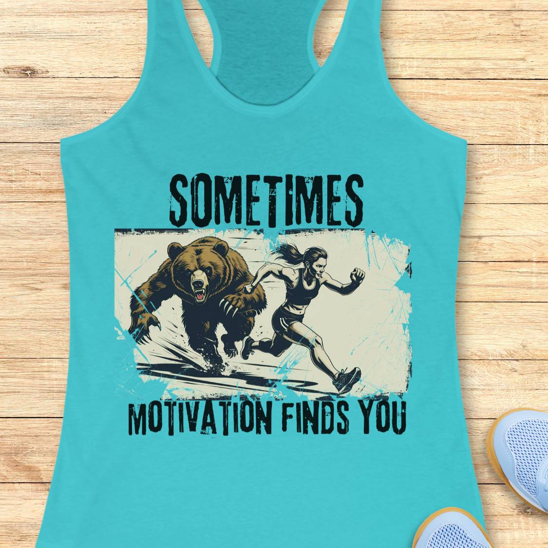 A turquoise racerback tank top laid flat on a light wood surface, featuring a distressed graphic of a bear chasing a woman running, with the text 'SOMETIMES MOTIVATION FINDS YOU' in black. Part of a light blue shoe is visible at the bottom right.