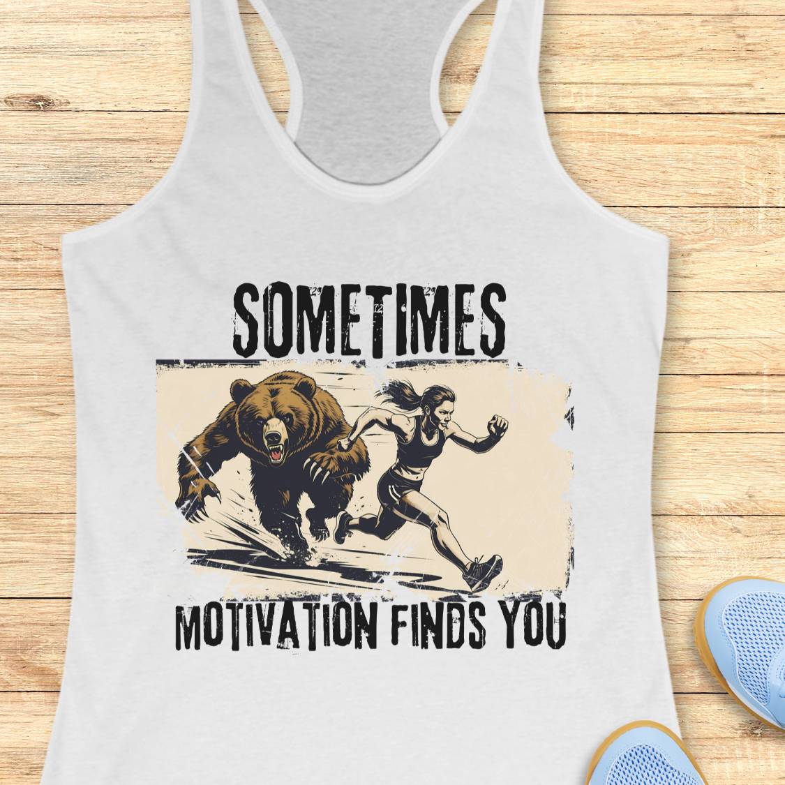 A white racerback tank top laid flat on a light wood surface, featuring a distressed graphic of a woman running from a bear with the text 'SOMETIMES MOTIVATION FINDS YOU.' A partial blue shoe is visible at the lower right as a styling prop.