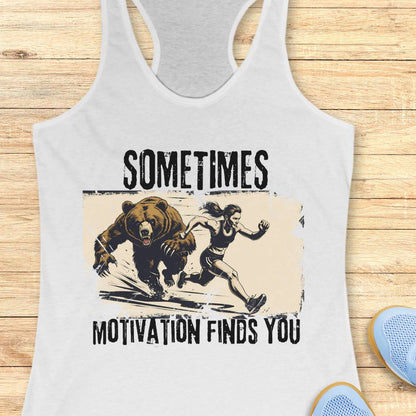 A white racerback tank top laid flat on a light wood surface, featuring a distressed graphic of a woman running from a bear with the text 'SOMETIMES MOTIVATION FINDS YOU.' A partial blue shoe is visible at the lower right as a styling prop.