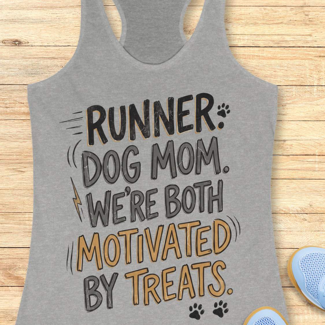 A heather gray racerback tank top laid flat on a light wood background, featuring the printed phrase 'RUNNER DOG MOM WE'RE BOTH MOTIVATED BY TREATS.' in black and mustard yellow lettering with small paw print and lightning bolt graphics.