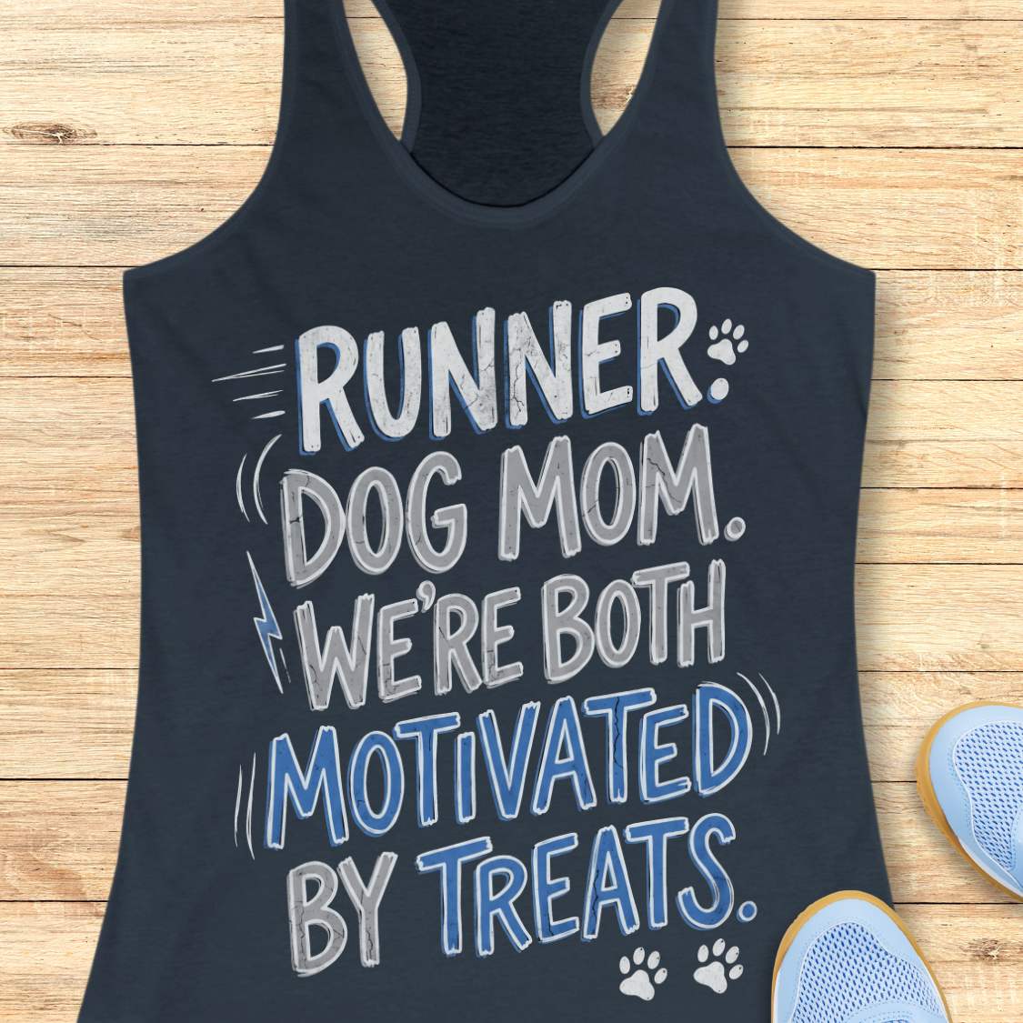A indigo racerback tank top laid flat on a light wood background, featuring the text 'RUNNER DOG MOM WE'RE BOTH MOTIVATED BY TREATS.' in white and blue lettering with small paw print graphics.