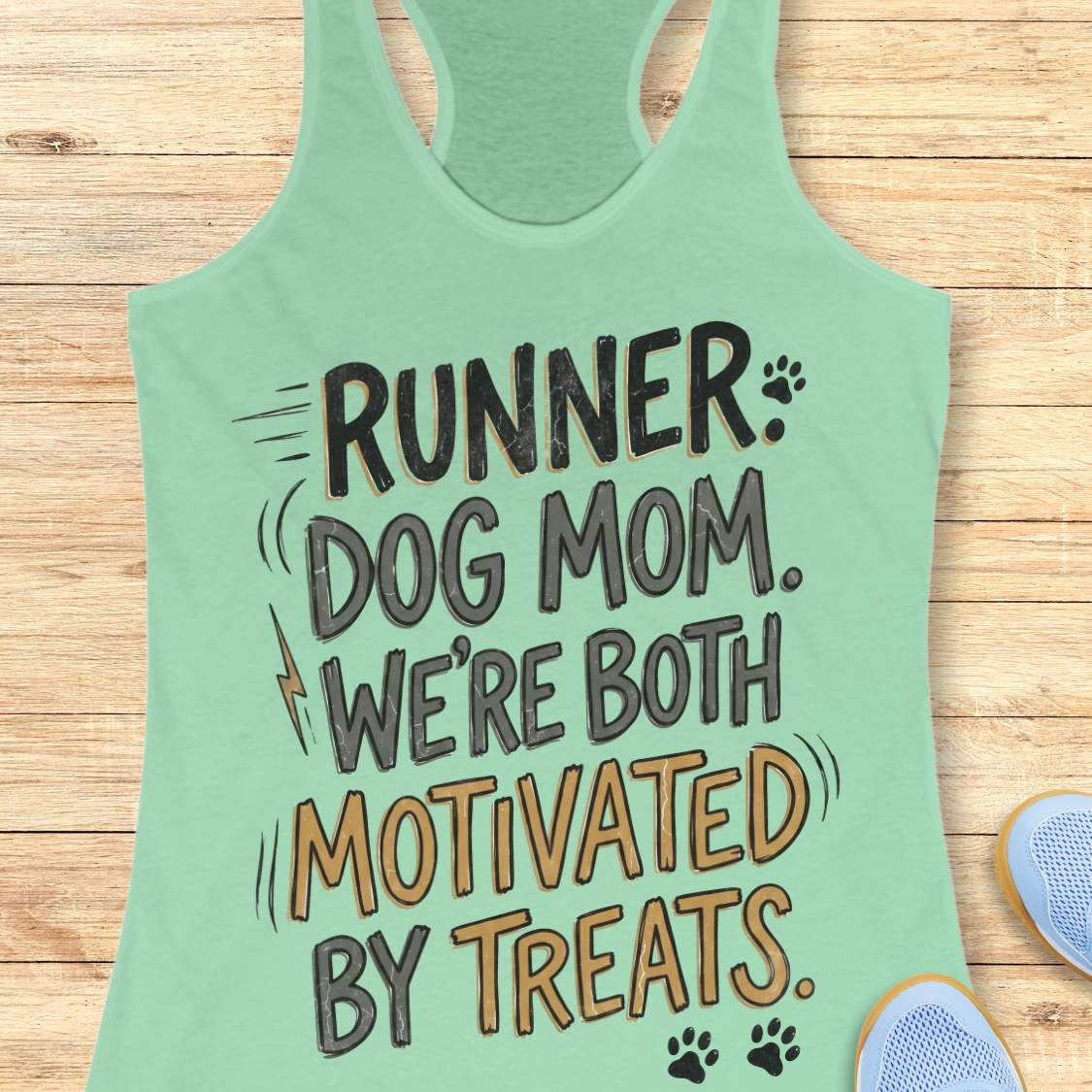 A mint racerback tank top laid flat on a light wood background, featuring the printed phrase 'RUNNER DOG MOM WE're BOTH MOTIVATED BY TREATS' in black and mustard yellow lettering with small paw print and lightning bolt accents. Partial blue shoes are visible at the bottom right as styling props.