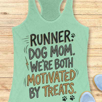 A mint racerback tank top laid flat on a light wood background, featuring the printed phrase 'RUNNER DOG MOM WE're BOTH MOTIVATED BY TREATS' in black and mustard yellow lettering with small paw print and lightning bolt accents. Partial blue shoes are visible at the bottom right as styling props.