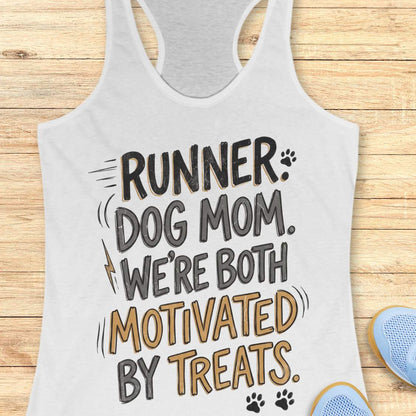 A white racerback tank top laid flat on a light wood background, featuring the printed phrase 'RUNNER DOG MOM WE BOTH MOTIVATED BY TREATS' in black and mustard yellow lettering with small paw print and lightning bolt accents. Partial blue shoes are visible at the bottom right as styling props.