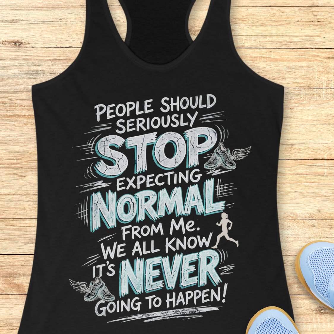 A black racerback tank top laid flat on a light wood background, featuring a large white and light blue graphic text that reads, 'People should seriously stop expecting normal from me. We all know it's never going to happen!' with small winged shoe illustrations and a running figure.
