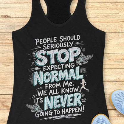 A black racerback tank top laid flat on a light wood background, featuring a large white and light blue graphic text that reads, 'People should seriously stop expecting normal from me. We all know it's never going to happen!' with small winged shoe illustrations and a running figure.