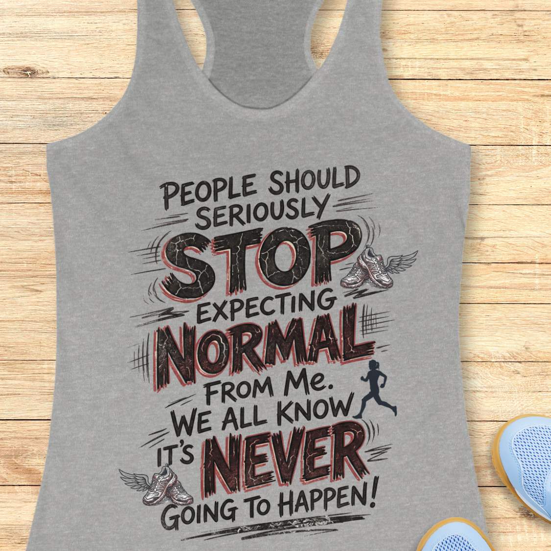 A heather gray racerback tank top with a motivational graphic print, displayed flat on a light wood background with blue sneakers partially visible in the corner.