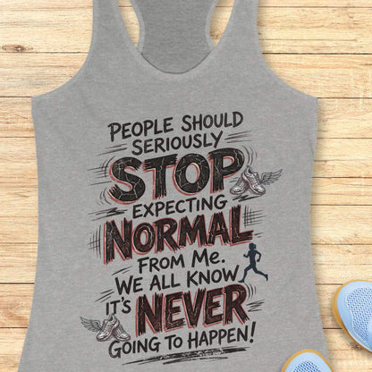 A heather gray racerback tank top with a motivational graphic print, displayed flat on a light wood background with blue sneakers partially visible in the corner.