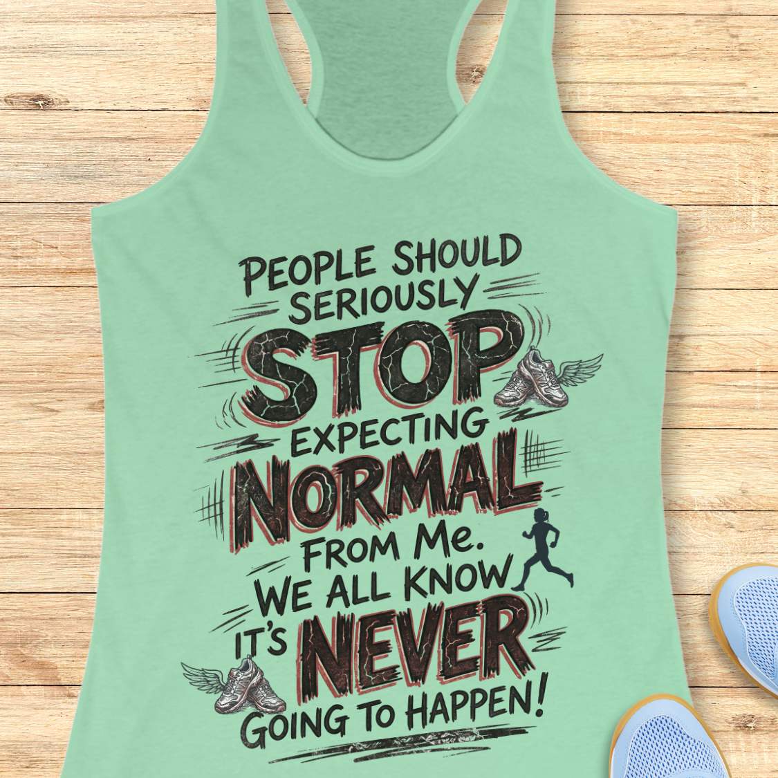A mint green racerback tank top with a humorous motivational graphic printed on the front, laid flat on a light wood background with partial sneakers visible at the bottom right.