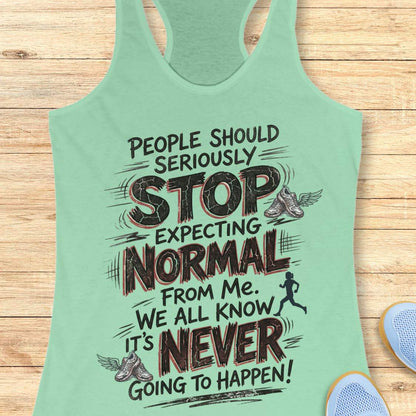 A mint green racerback tank top with a humorous motivational graphic printed on the front, laid flat on a light wood background with partial sneakers visible at the bottom right.