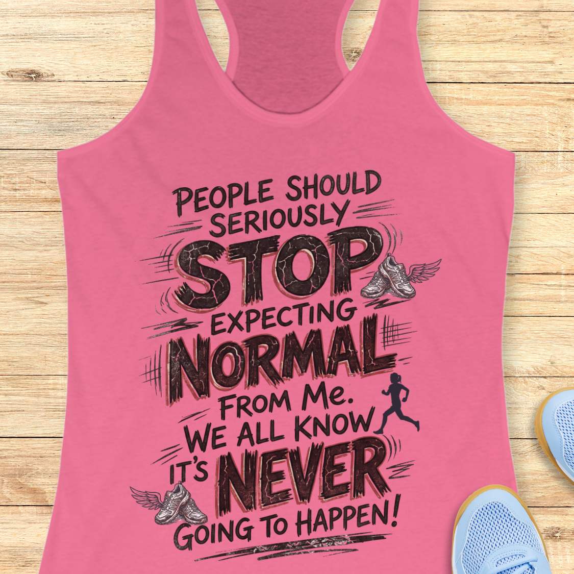 A pink racerback tank top with a motivational running quote printed in black text, displayed flat on a light wood background with partial blue sneakers visible at the edges.