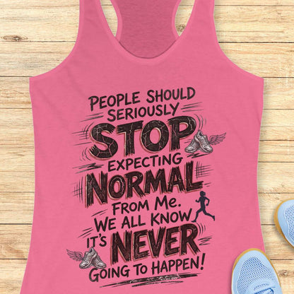 A pink racerback tank top with a motivational running quote printed in black text, displayed flat on a light wood background with partial blue sneakers visible at the edges.