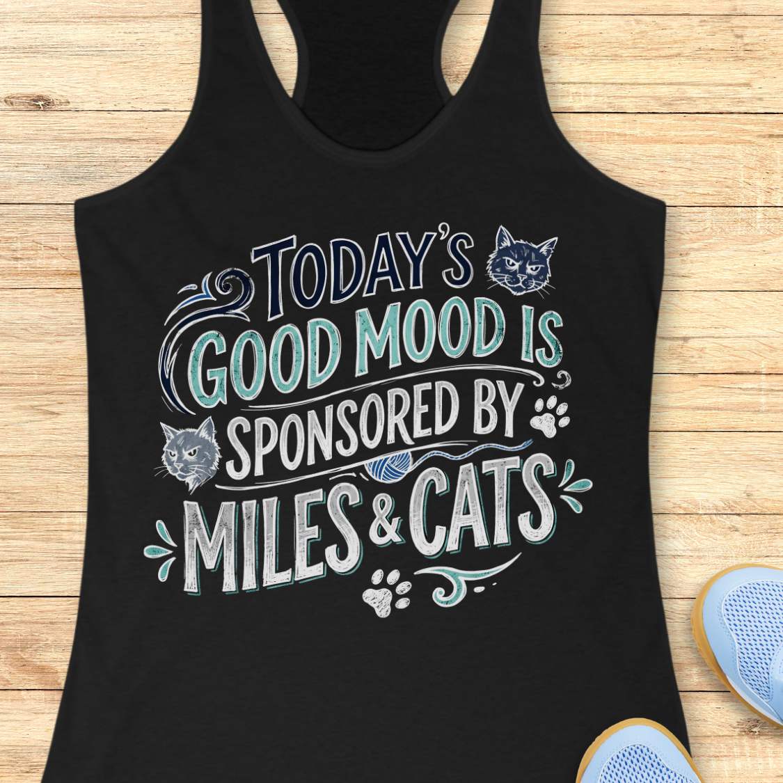 A black racerback tank top laid flat on a light wood background, featuring a stylized graphic that reads 'TODAY'S GOOD MOOD IS SPONSORED BY MILES & CATS' in white and teal lettering with cat illustrations and paw prints. Part of a blue shoe is visible at the bottom right.