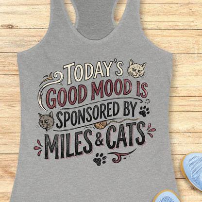 A heather gray racerback tank top laid flat on a light wood background, featuring a humorous graphic that reads 'TODAY'S GOOD MOOD IS SPONSORED BY MILES & CATS' with illustrations of two cats, paw prints, and a small ball of yarn.