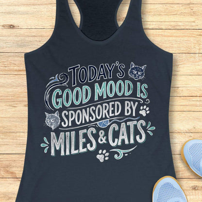A indigo racerback tank top laid flat on a light wood background, featuring a humorous graphic that reads 'TODAY'S GOOD MOOD IS SPONSORED BY MILES & CATS' in white and teal lettering with cat illustrations and paw prints. Part of a blue shoe is visible in the bottom right corner.