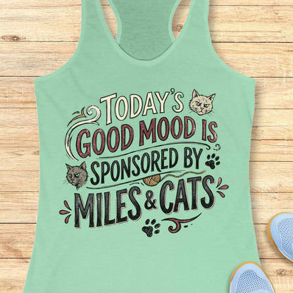 A mint green racerback tank top laid flat on a light wood background, featuring a humorous graphic that reads 'TODAY'S GOOD MOOD IS SPONSORED BY MILES & CATS' with illustrations of two cats and paw prints.