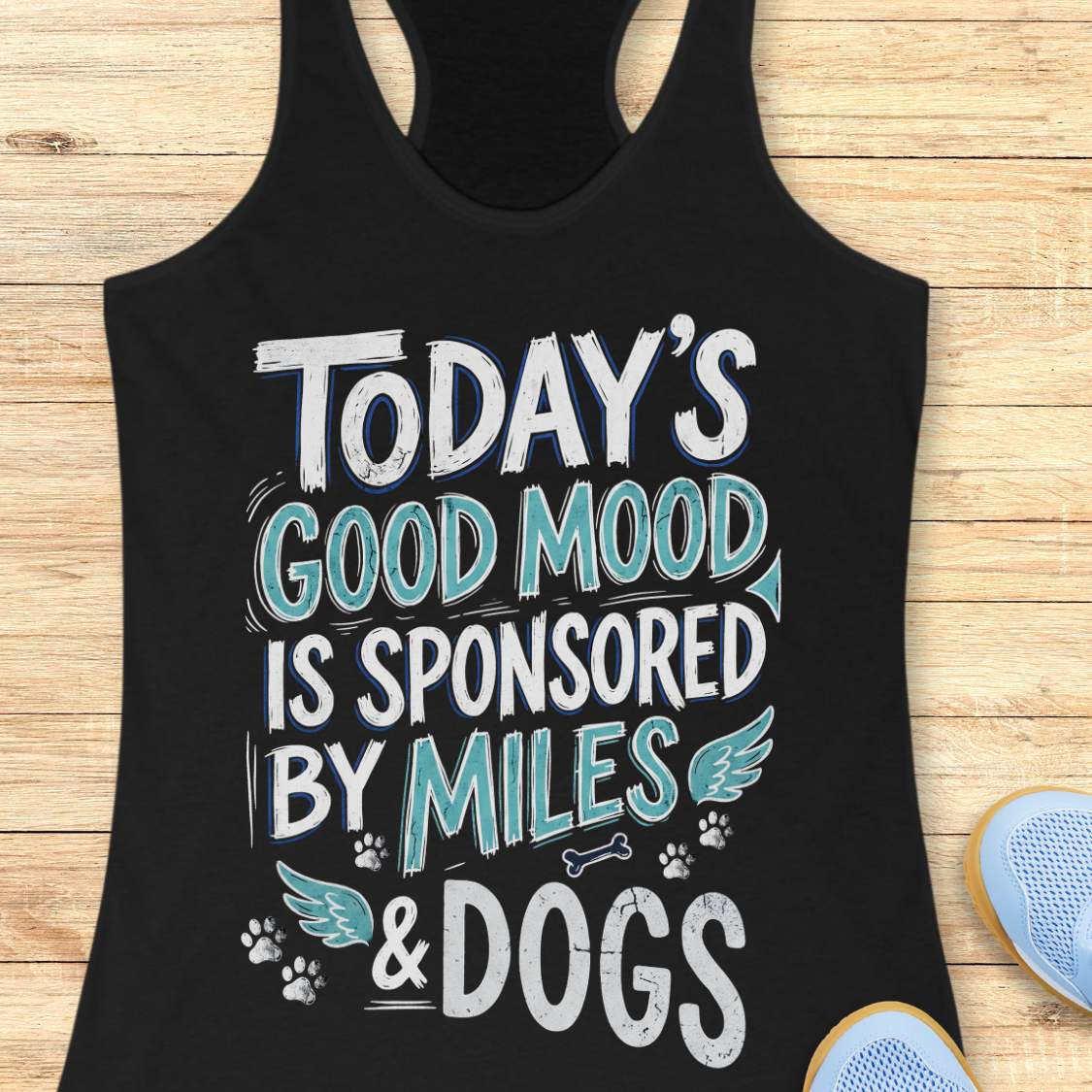 A black racerback tank top laid flat on a light wood background, featuring the text 'TODAY'S GOOD MOOD IS SPONSORED BY MILES & DOGS' in white and teal lettering with small dog-themed graphics. Part of a blue shoe is visible at the bottom right.