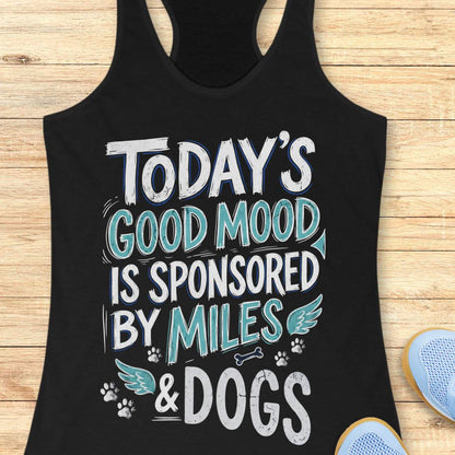 A black racerback tank top laid flat on a light wood background, featuring the text 'TODAY'S GOOD MOOD IS SPONSORED BY MILES & DOGS' in white and teal lettering with small dog-themed graphics. Part of a blue shoe is visible at the bottom right.