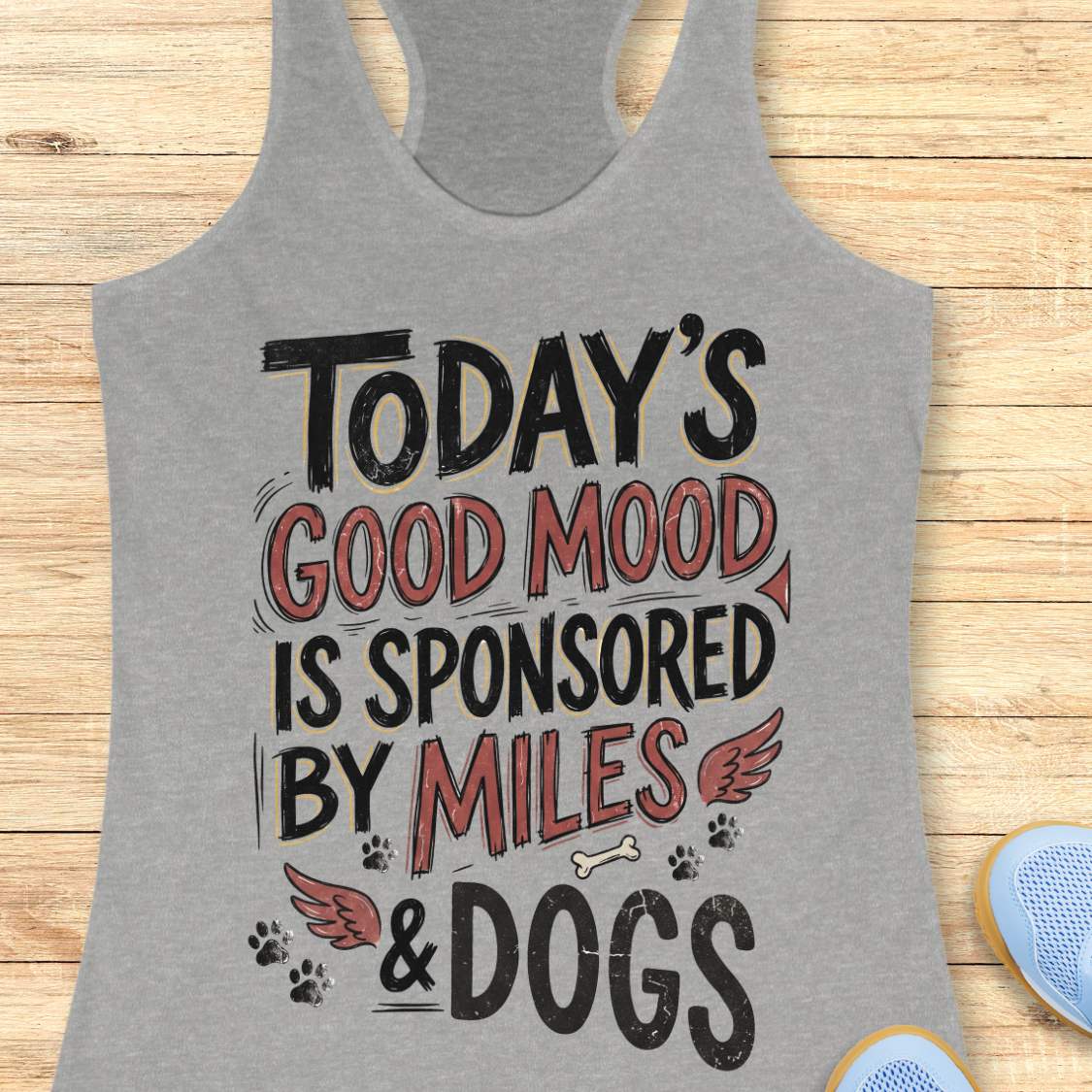 A heather gray racerback tank top laid flat on a light wood background, featuring a graphic print that reads 'TODAY'S GOOD MOOD IS SPONSORED BY MILES & DOGS' in black and muted red lettering with small dog-themed illustrations.