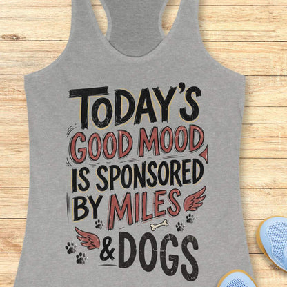A heather gray racerback tank top laid flat on a light wood background, featuring a graphic print that reads 'TODAY'S GOOD MOOD IS SPONSORED BY MILES & DOGS' in black and muted red lettering with small dog-themed illustrations.